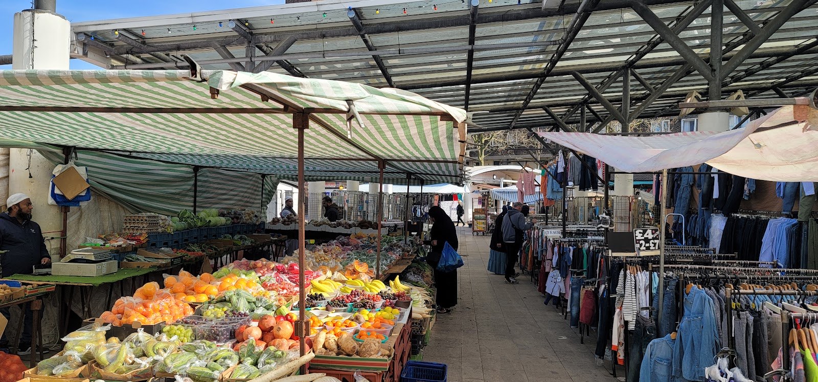Shoppers browsing fruit, vegetable and clothing stalls under the steel canopy at Chrisp Street Market, Poplar E14